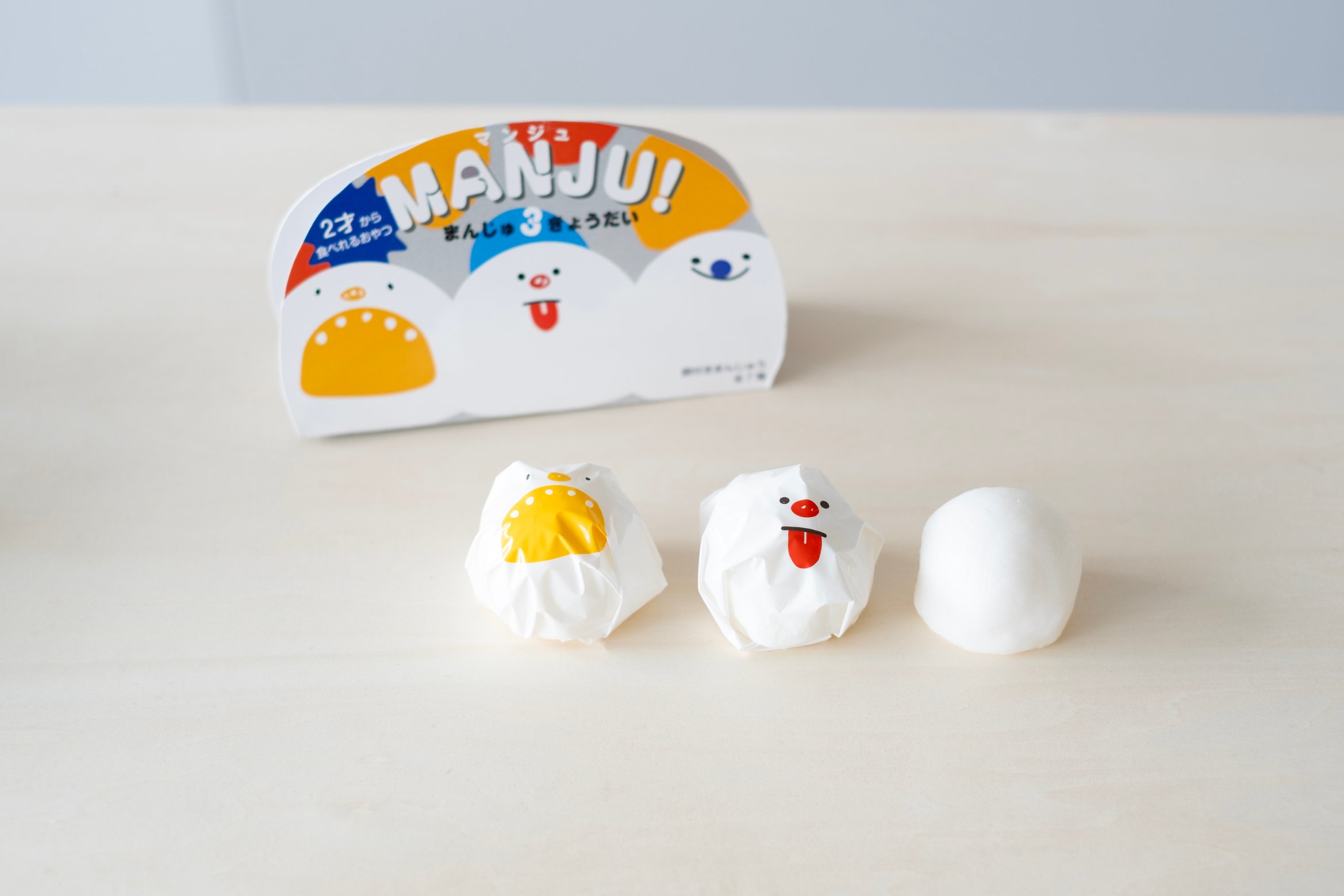 まんじゅうを顔が印刷された包装紙で包んでいる。外を包むパッケージに「MANJU!」「まんじゅ3きょうだい」と書かれている。