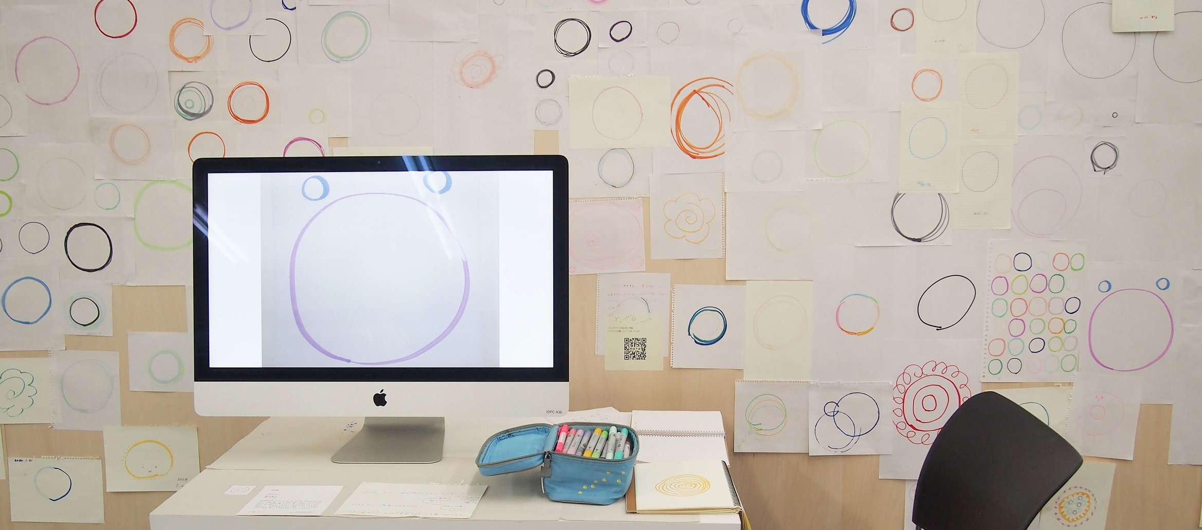 iMacの後に壁一面を覆う大きな木の板に丸が描かれた紙が沢山貼られている。