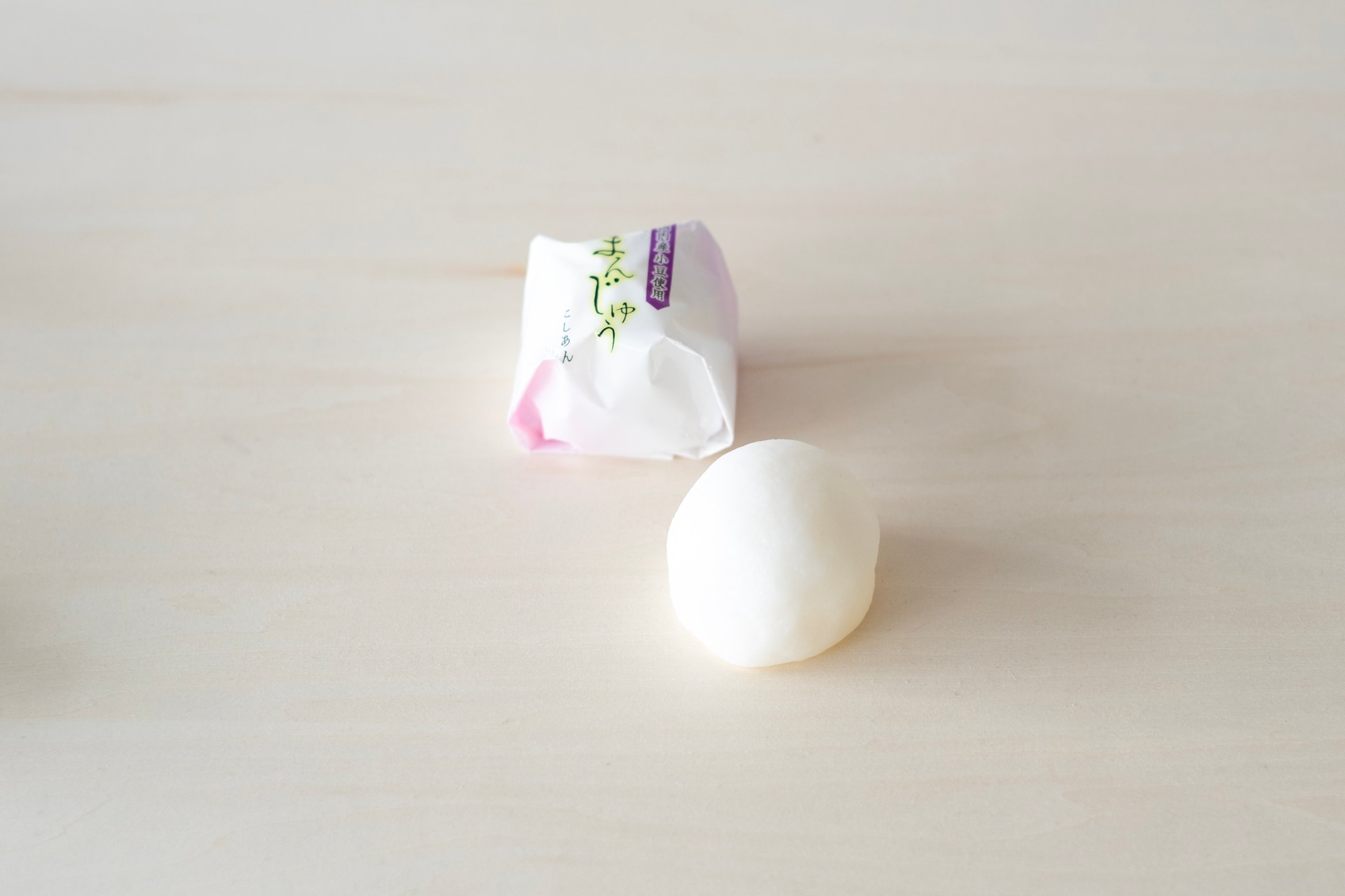 まんじゅうが白色と薄ピンク色のグラデーションの包装紙に包まれている。包装紙には「まんじゅう」と達筆な字で縦書きに書かれている。